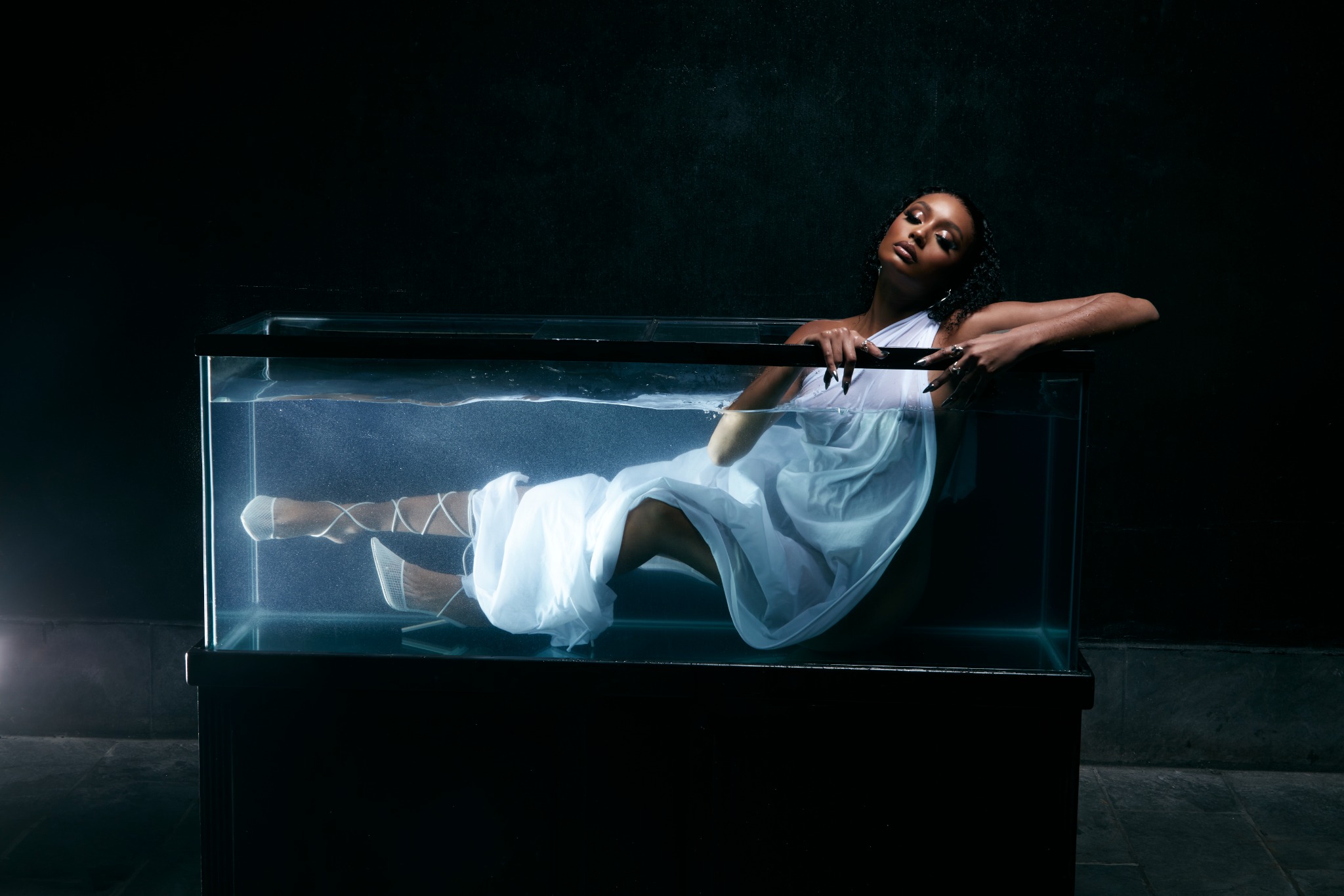 A fashion portrait of a woman in white reclining in an illuminated glass tank